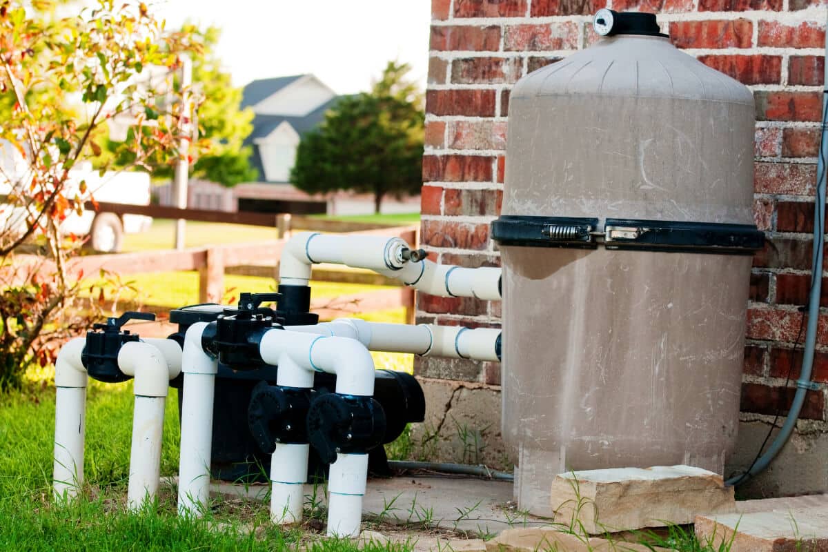 A sand filter tank and PVC valve manifold installed beside a brick house